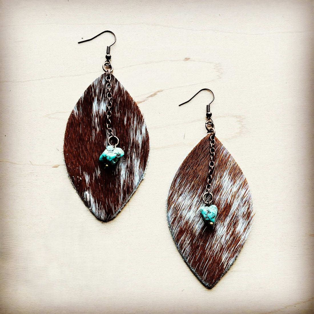 A pair of oval-shaped hair-on-hide leather earrings with brown and white speckled fur and a turquoise stone drop.