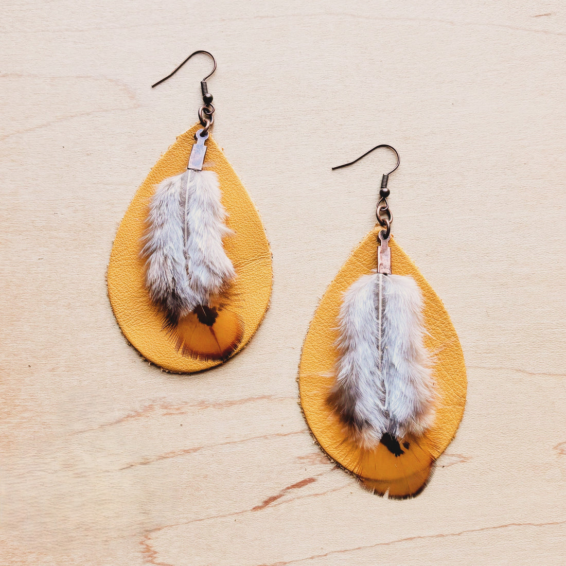 Mustard yellow leather teardrop earrings with soft feather details hanging on a light wood background