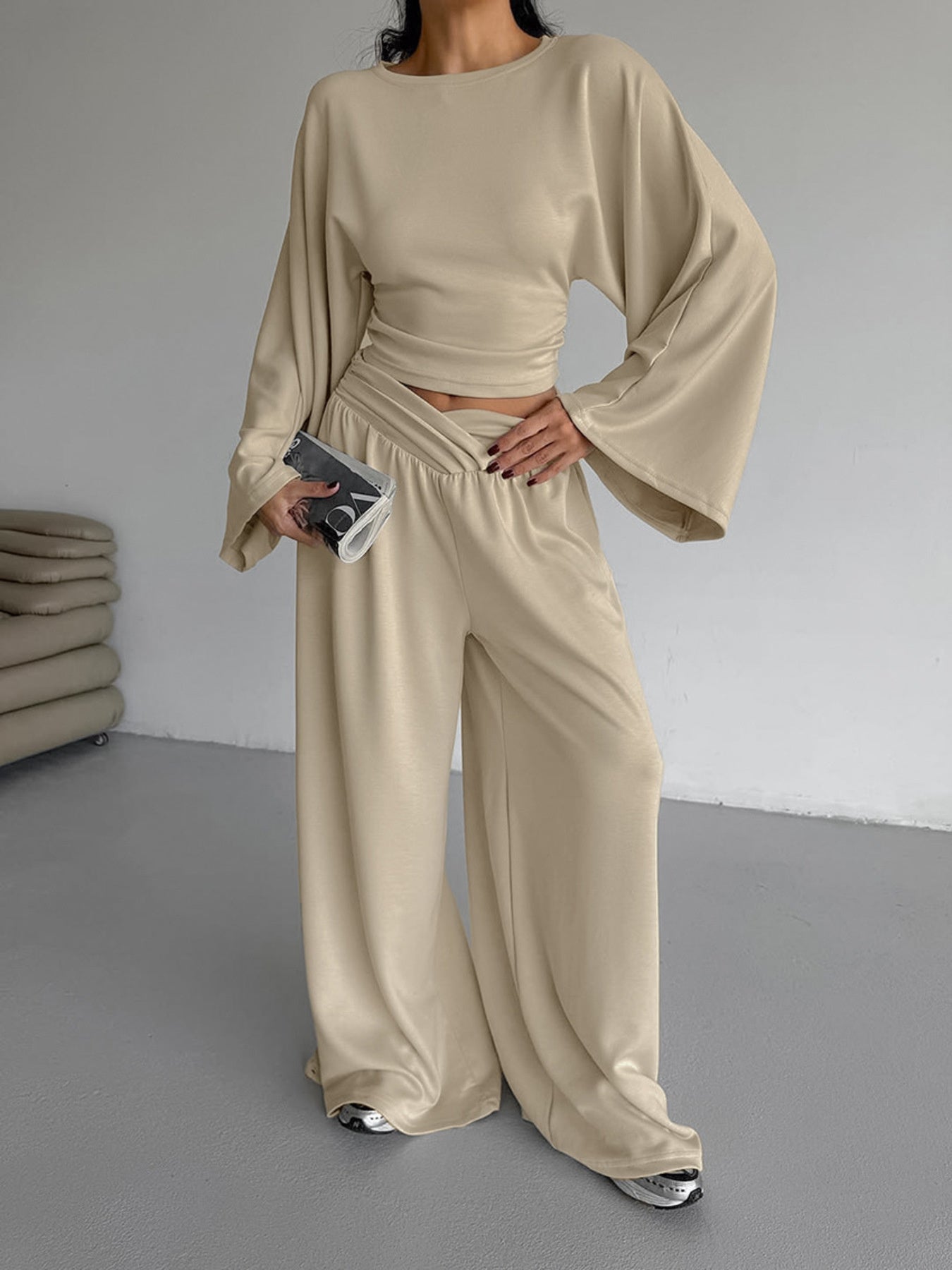 Person wearing a beige outfit with wide sleeves and pants, holding a phone.