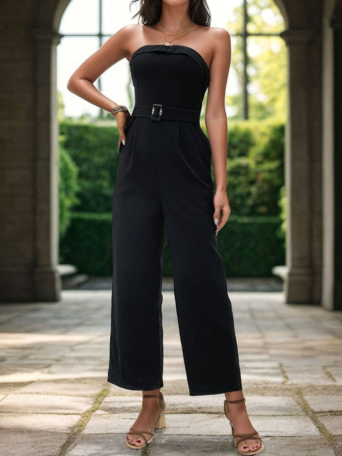A woman wearing a black strapless tube jumpsuit with a waist belt and wide legs, standing in an outdoor setting.