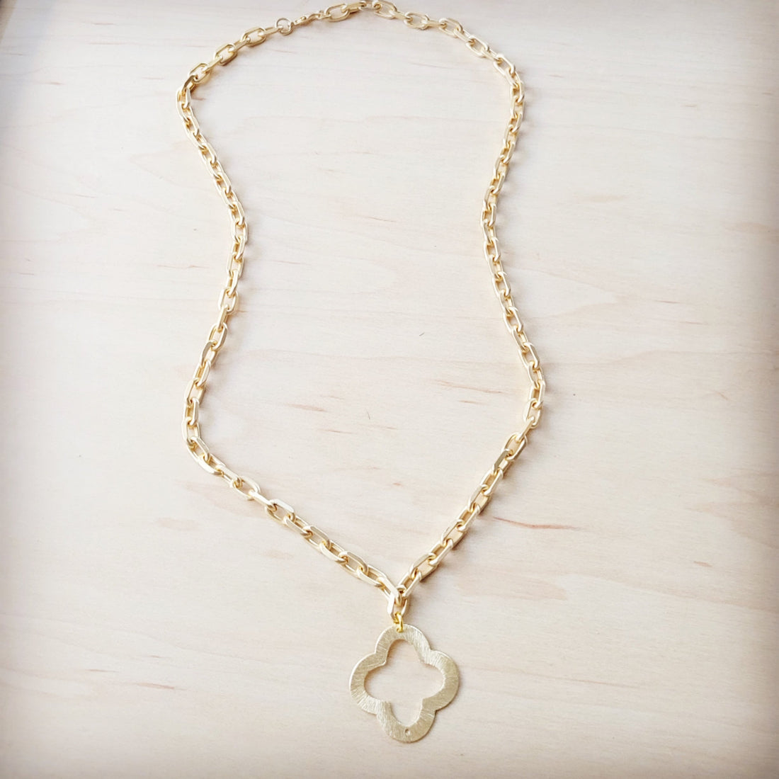 Matte gold quatrefoil pendant hanging on a matching gold-tone chain against a light wood background.