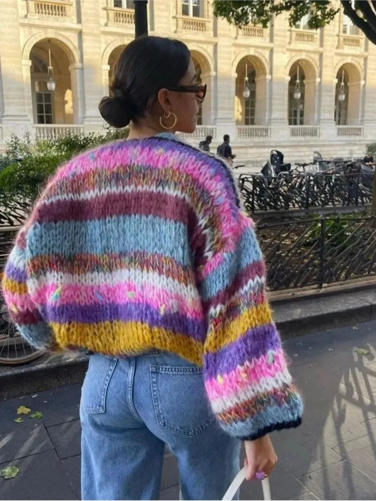 A woman wearing a vibrant multi-colored striped crochet cardigan over a white crop top and blue jeans, standing outdoors.
