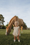 Baby wearing a cream muslin romper with a brown horse print standing in a grassy field with a horse in the background