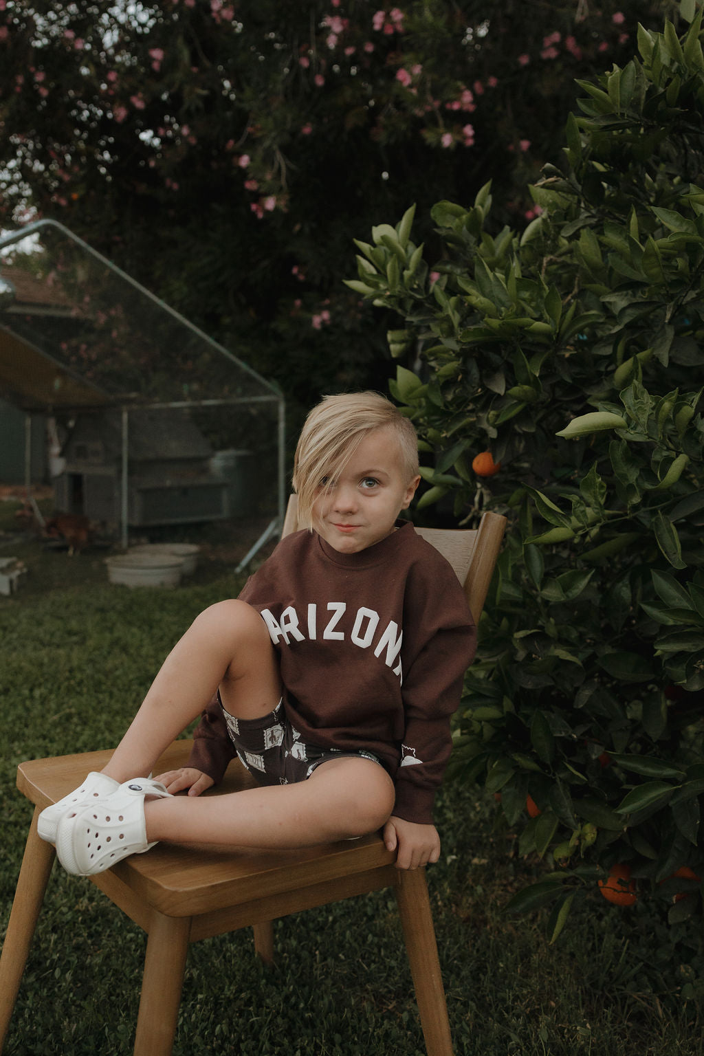 A child wearing a brown long-sleeve Arizona graphic sweatshirt while sitting on a wooden chair.