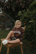 A child wearing a brown long-sleeve Arizona graphic sweatshirt while sitting on a wooden chair.