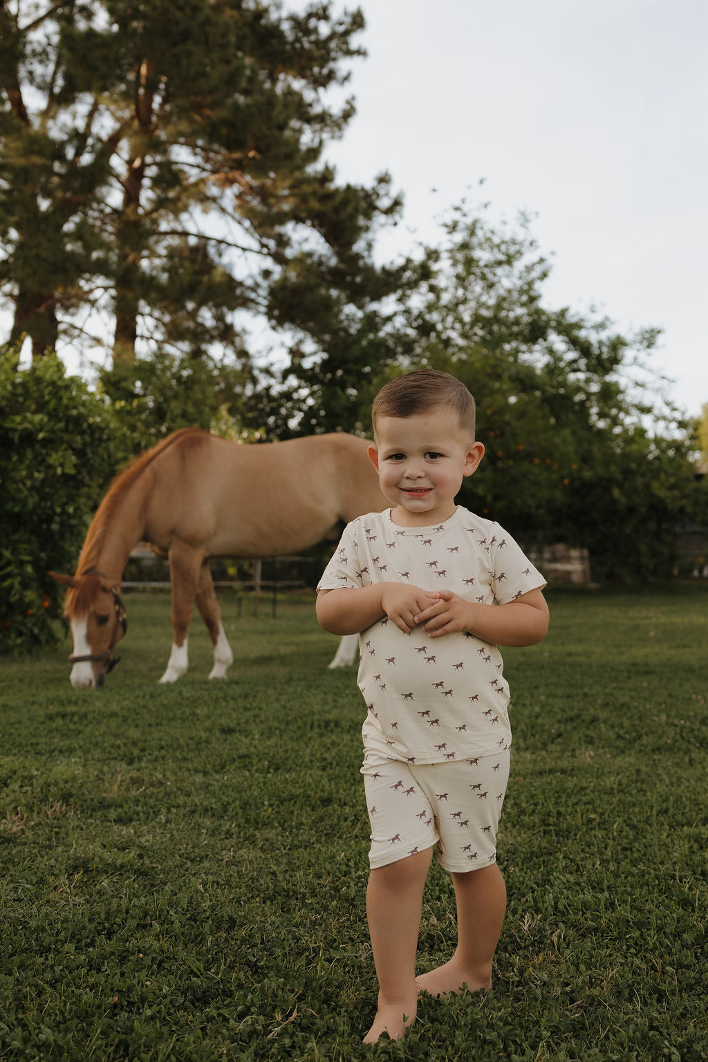 A young boy wearing a cream colored bamboo short set with a wild horse print, standing in a grass field.