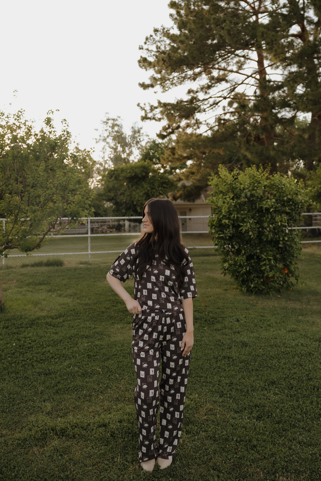 A woman wearing a brown lounge set featuring a With Love from AZ graphic print, consisting of a short-sleeve boxy tee and wide-leg pants, standing in a grassy area.