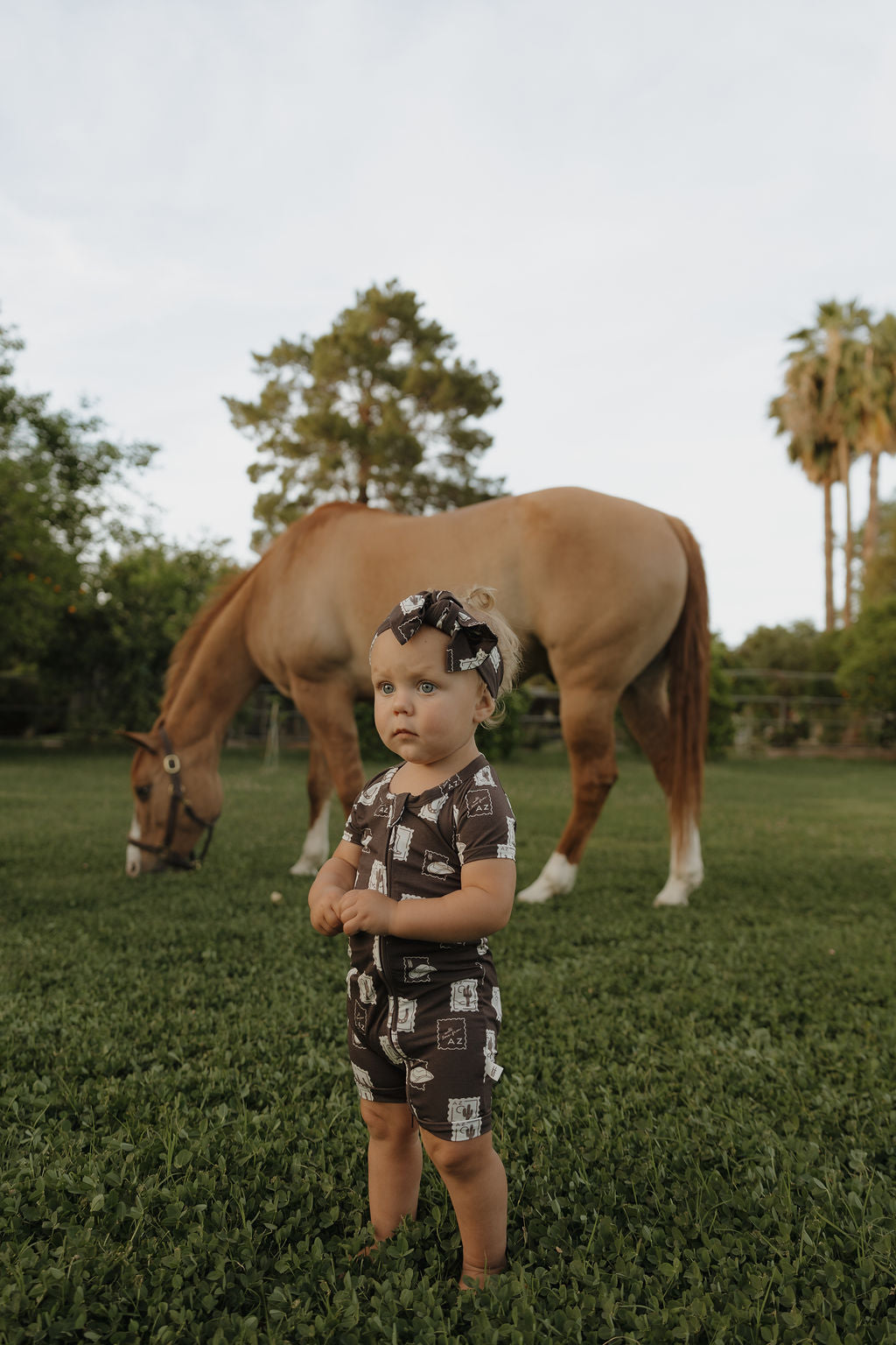 Baby wearing a brown shortie romper with desert-themed stamps and a matching headband standing in a grassy field.