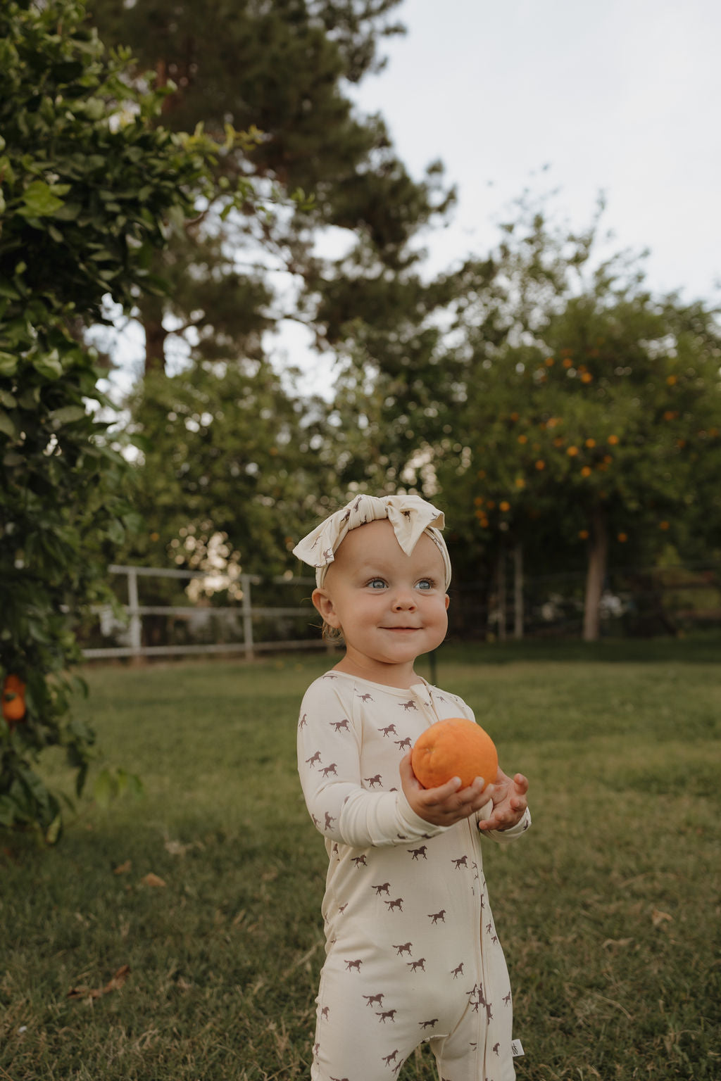 Baby wearing wild horse print bamboo zip pajamas with matching headband in a garden setting