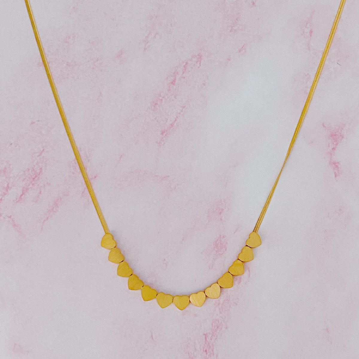 Gold chain necklace featuring a row of small heart charms on a pink marble background