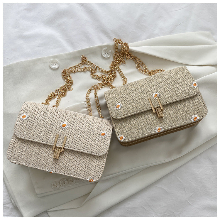 Two woven daisy chain crossbody bags in white and khaki, featuring gold-tone chain straps and a front clasp, resting on a white jacket.