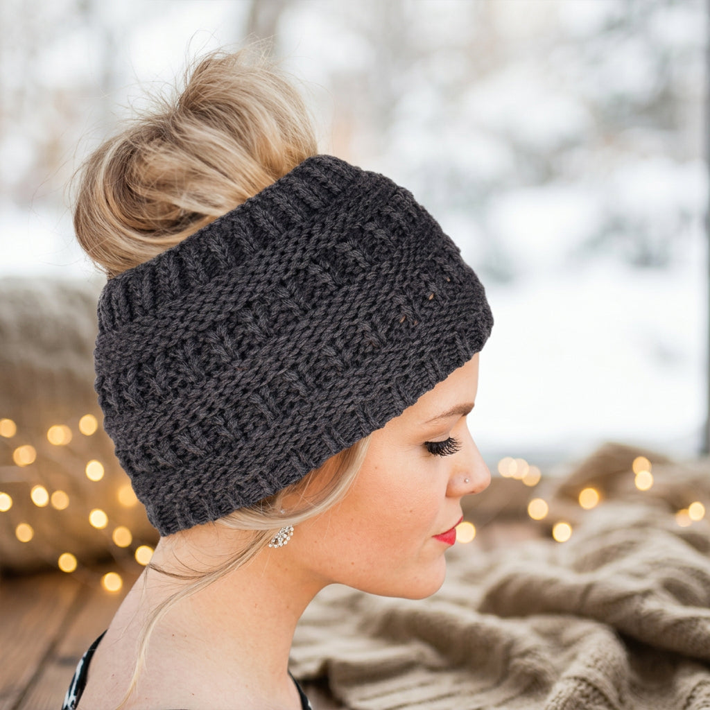 A woman in profile wearing a dark charcoal gray chunky knit beanie with a bun hairstyle, set against a cozy, softly blurred background with warm bokeh lights.