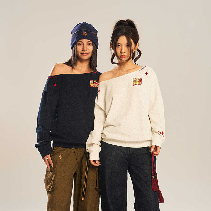 A woman wearing a beige long-sleeve sweatshirt with an asymmetric slanted shoulder and colorful embroidery details.
