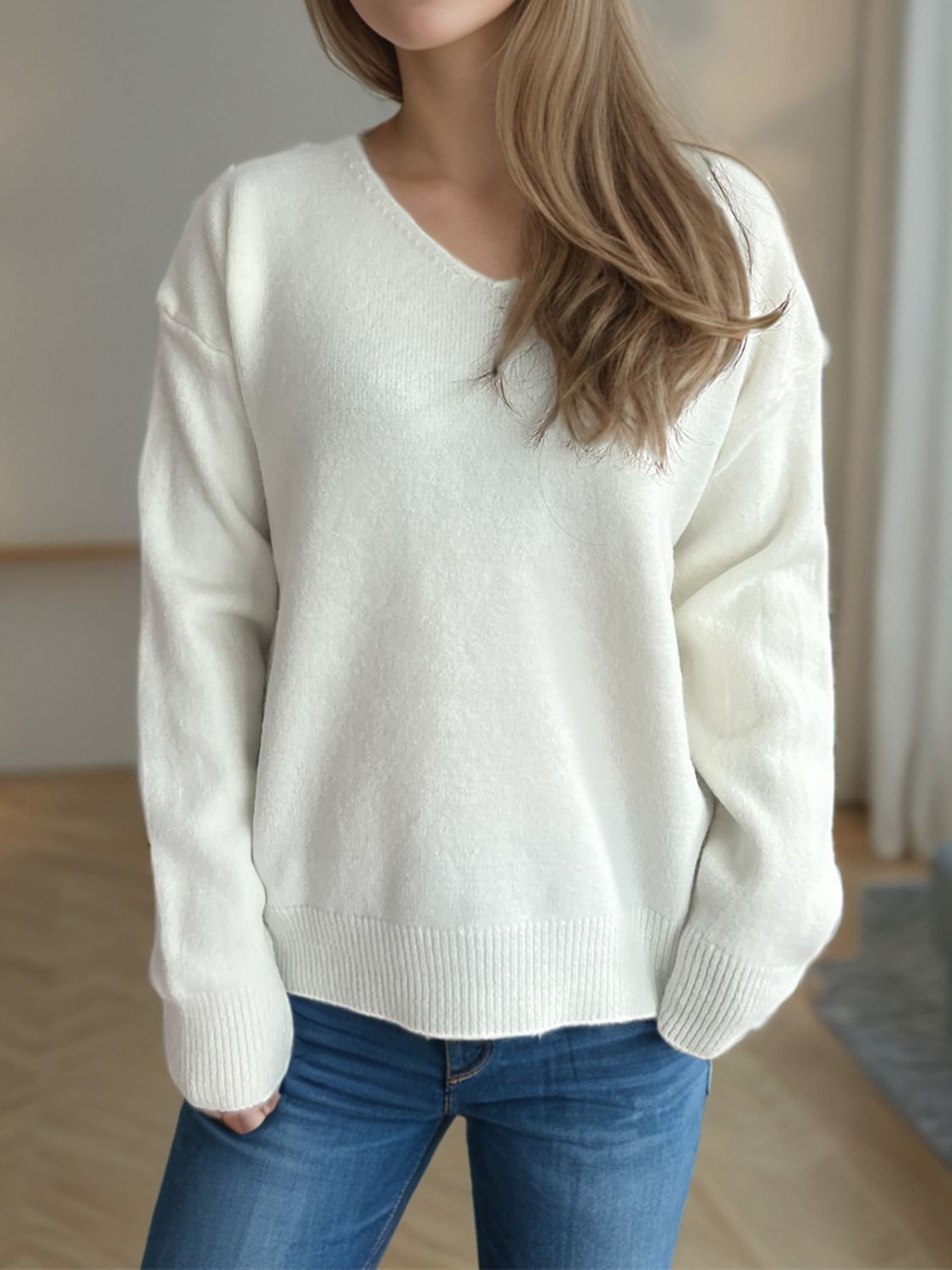 Woman wearing a white V-neck long sleeve sweater with dropped shoulders, paired with blue jeans.