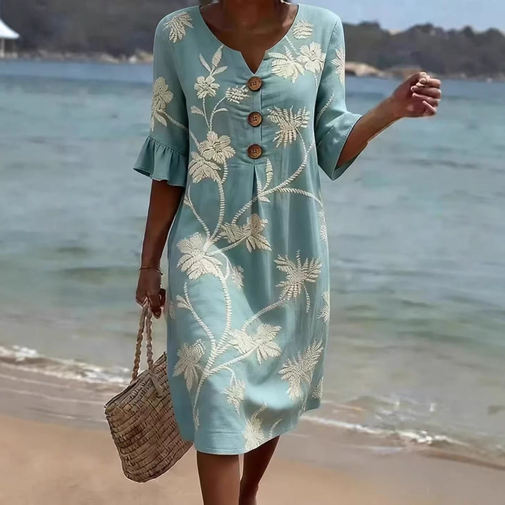 A woman wearing a light blue midi dress with a white palm tree print, v-neck, button details, and ruffled three-quarter sleeves on a beach.