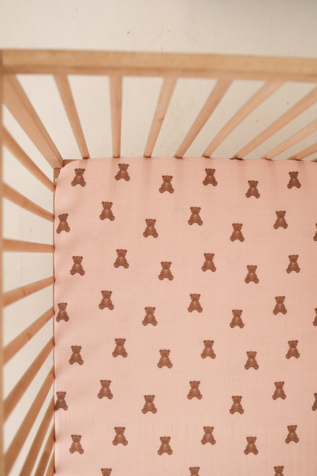 Close up of a light pink muslin baby crib sheet with a repeating brown bear print inside a wooden crib.