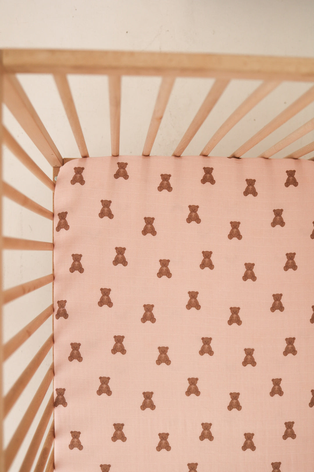 Close up of a light pink muslin baby crib sheet with a repeating brown bear print inside a wooden crib.