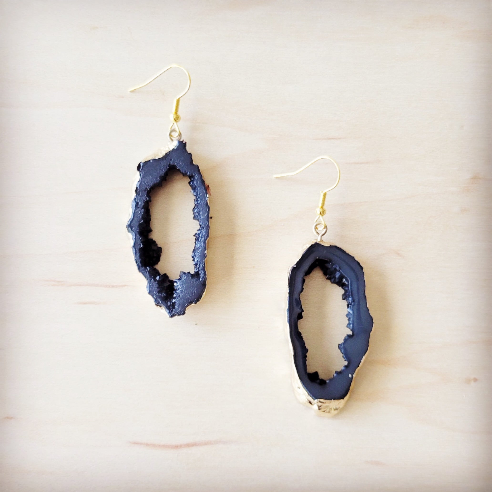 A pair of black crystalized geode dangle earrings with gold-tone edges on a light wooden background.
