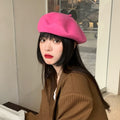 A vibrant pink wool blend beret worn by a model, and an olive green wool blend beret shown flat lay with a book, highlighting the candy-colored, retro Harajuku style.