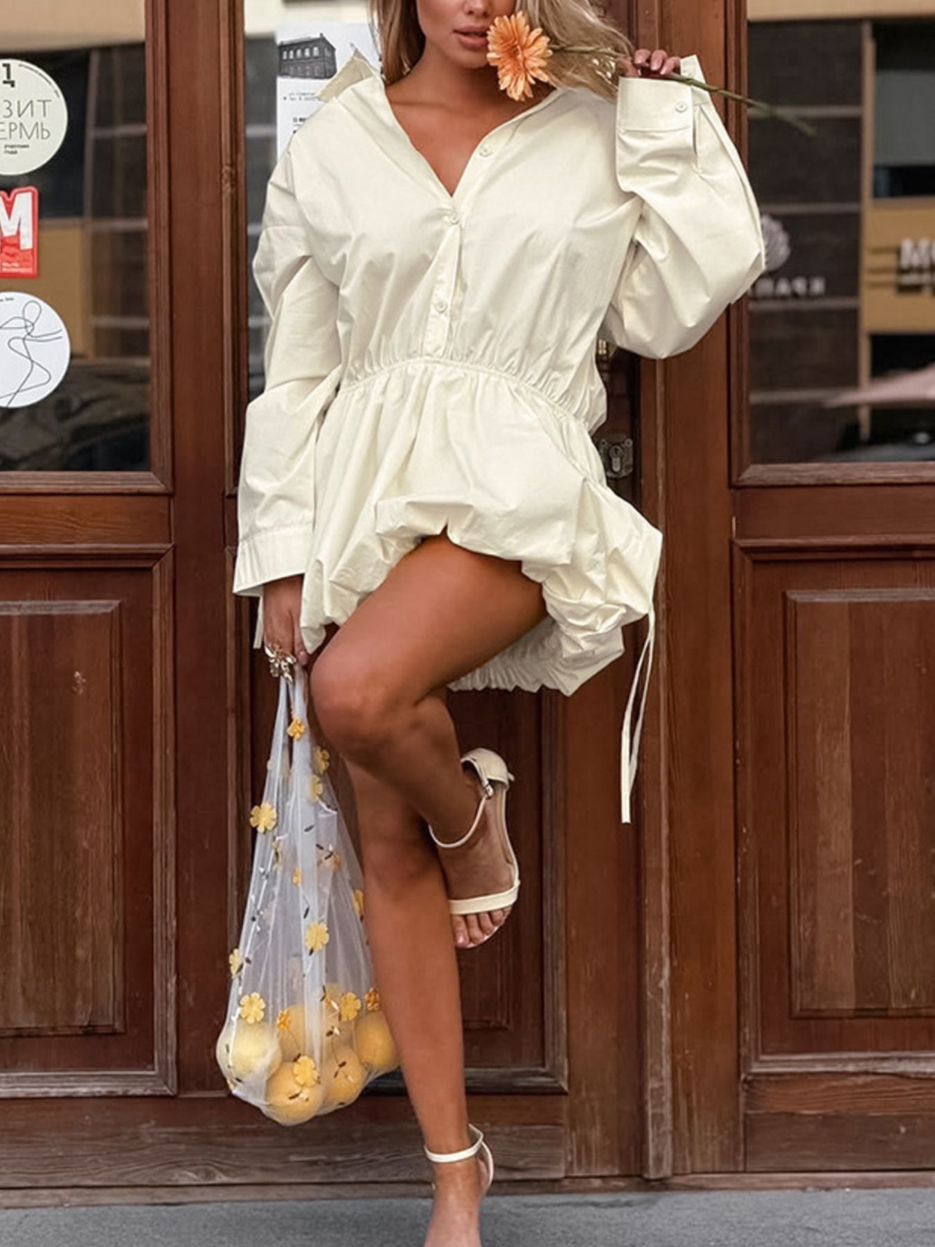 Woman in a white dress holding a flower, standing in front of wooden doors.