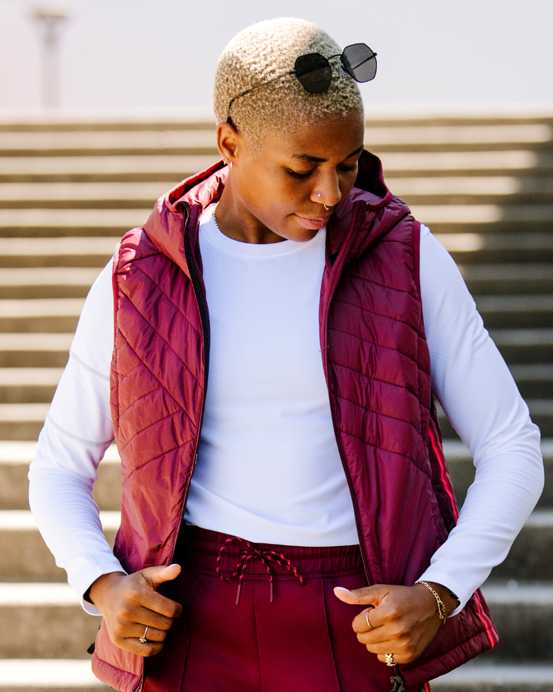 A person wearing a burgundy Powerstripe quilted hooded vest over a long sleeve white top, standing on concrete stairs.