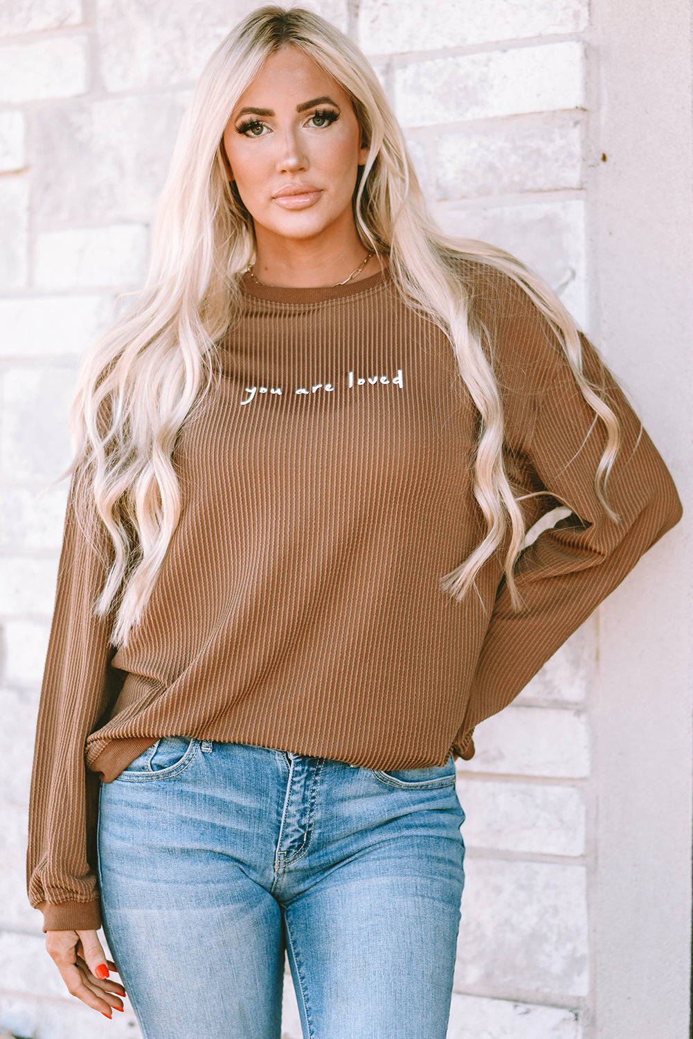 A woman wearing a brown ribbed long sleeve sweatshirt with the words You Are Loved printed on the chest.