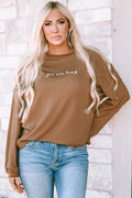 A woman wearing a brown ribbed long sleeve sweatshirt with the words You Are Loved printed on the chest.