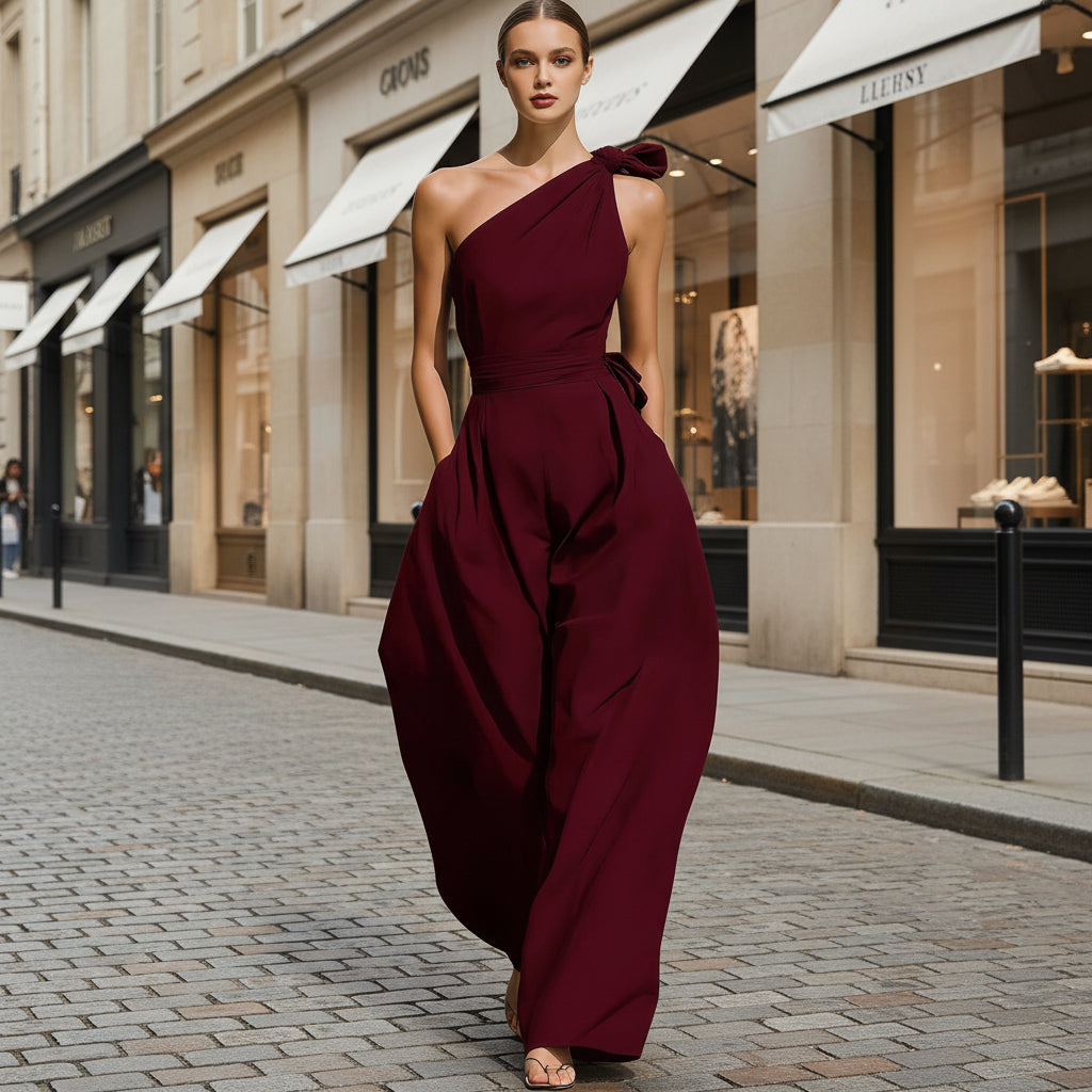 A woman wearing a deep maroon one-shoulder jumpsuit with a cinched waist and wide legs on a city street.