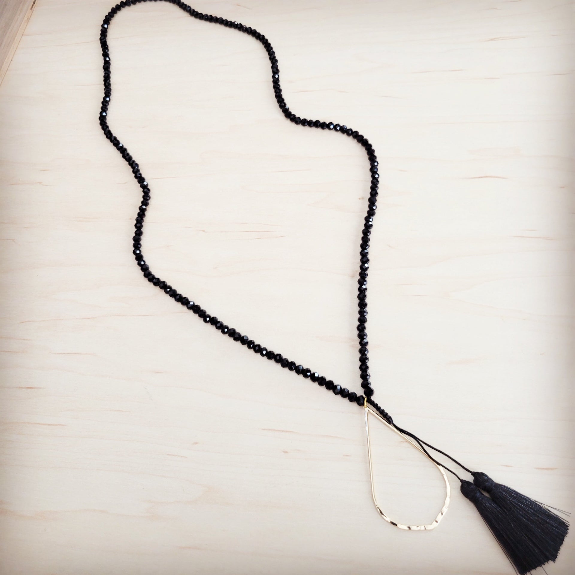 A black faceted bead lariat necklace with a gold-tone hammered teardrop charm and black fabric tassels.