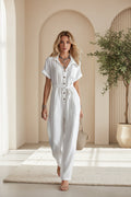 A woman wearing a crisp white loose button-down jumpsuit with a tie waist and side pockets in a bright, modern room.