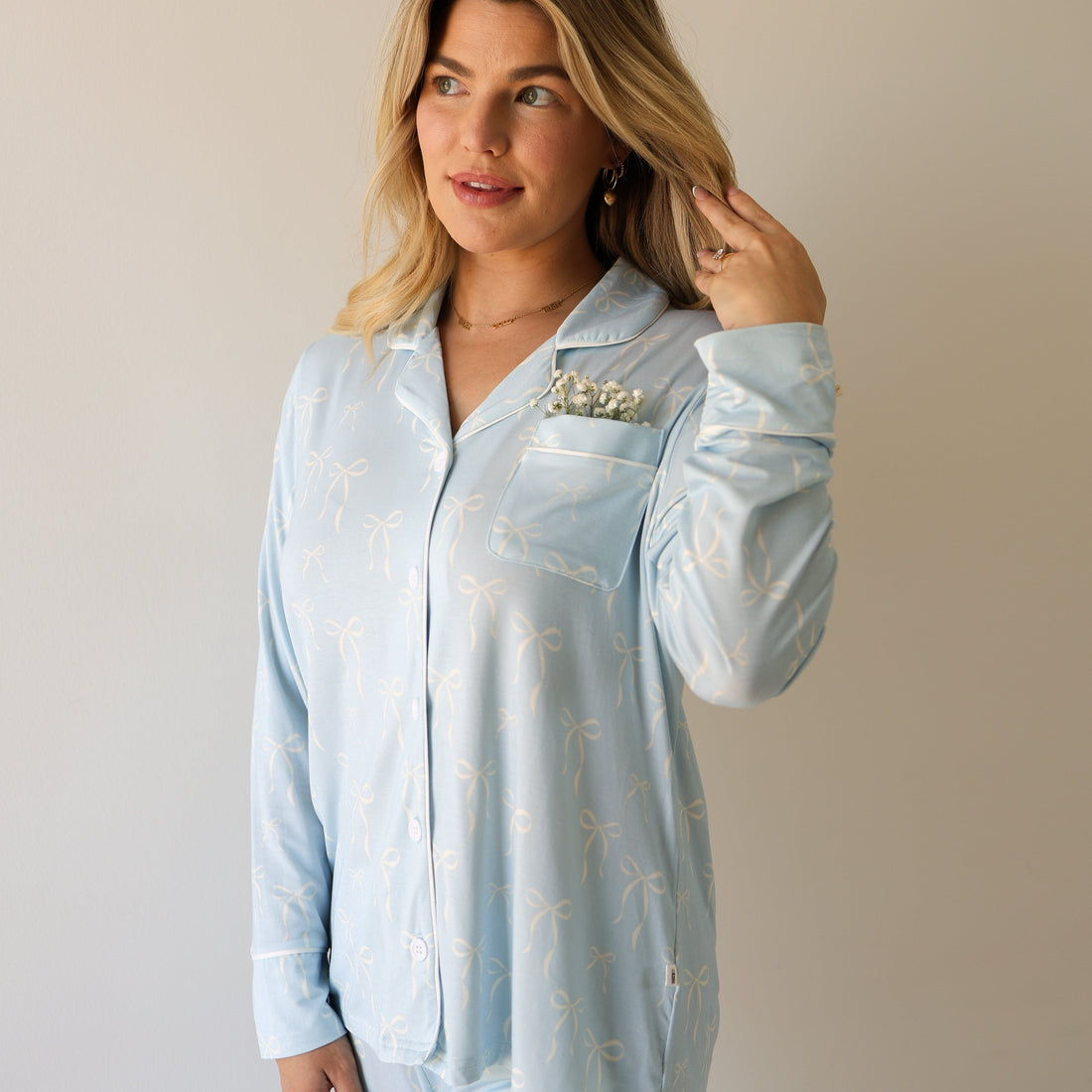 A woman wearing a light blue long-sleeve button-up pajama top with a white bow pattern, featuring a chest pocket with white piping.
