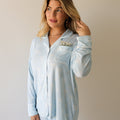 A woman wearing a light blue long-sleeve button-up pajama top with a white bow pattern, featuring a chest pocket with white piping.