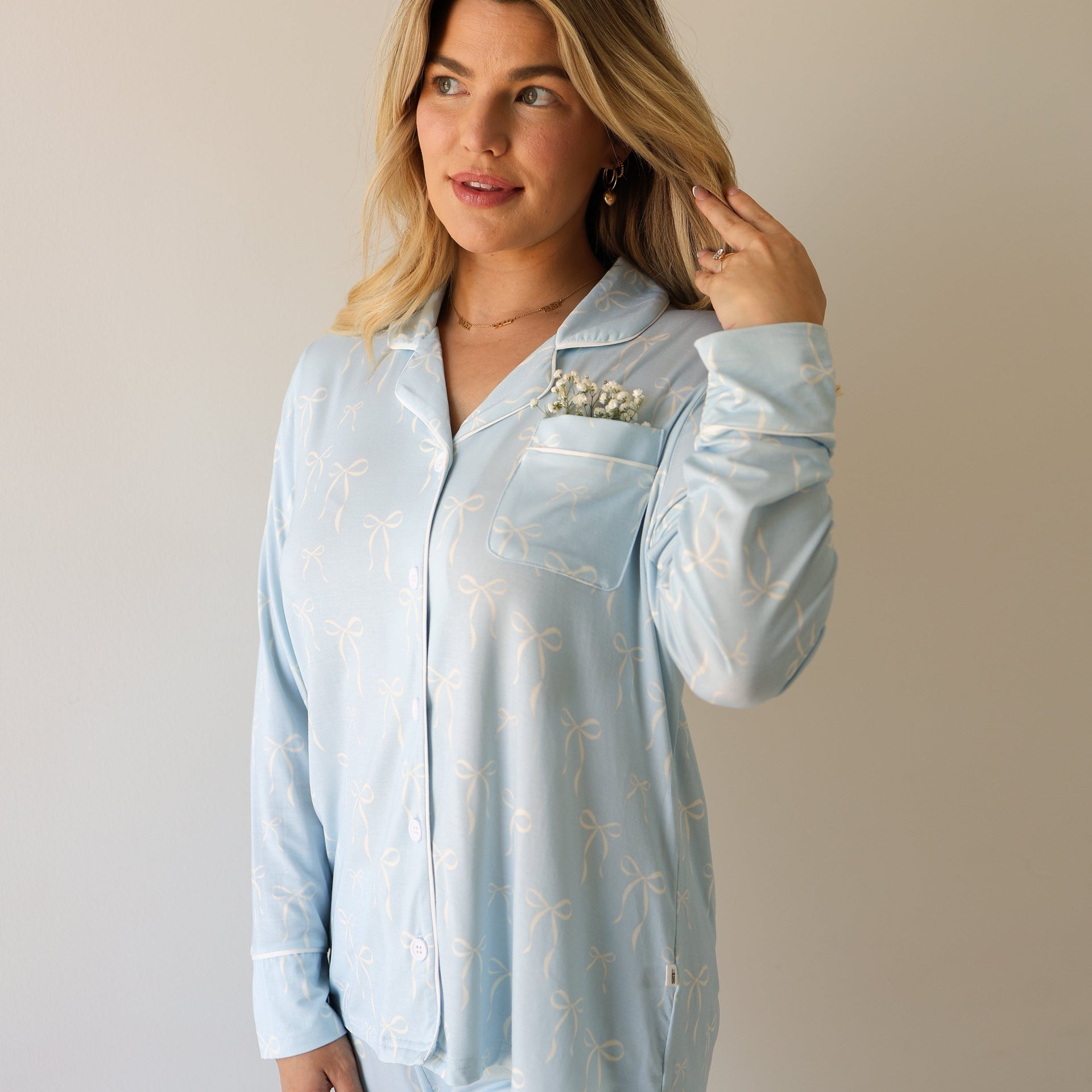 A woman wearing a light blue long-sleeve button-up pajama top with a white bow pattern, featuring a chest pocket with white piping.