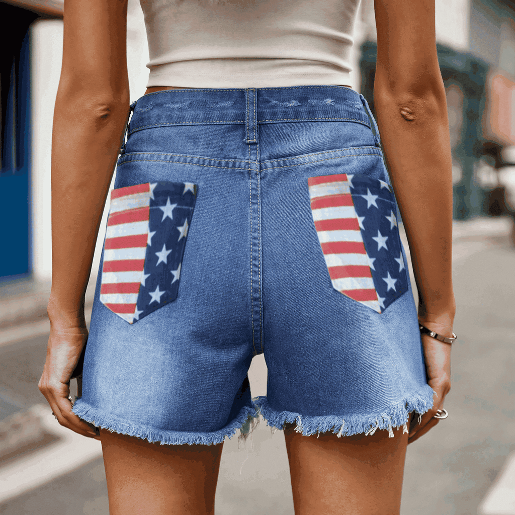 Close-up back view of blue denim shorts with US flag design on back pockets and frayed hem details.