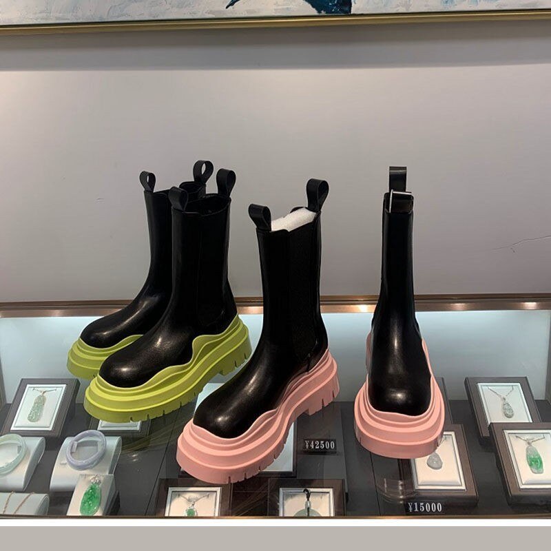 Pair of black mid-calf platform boots with a chunky neon green wavy sole, displayed next to a pair with a light pink sole, on a retail display.