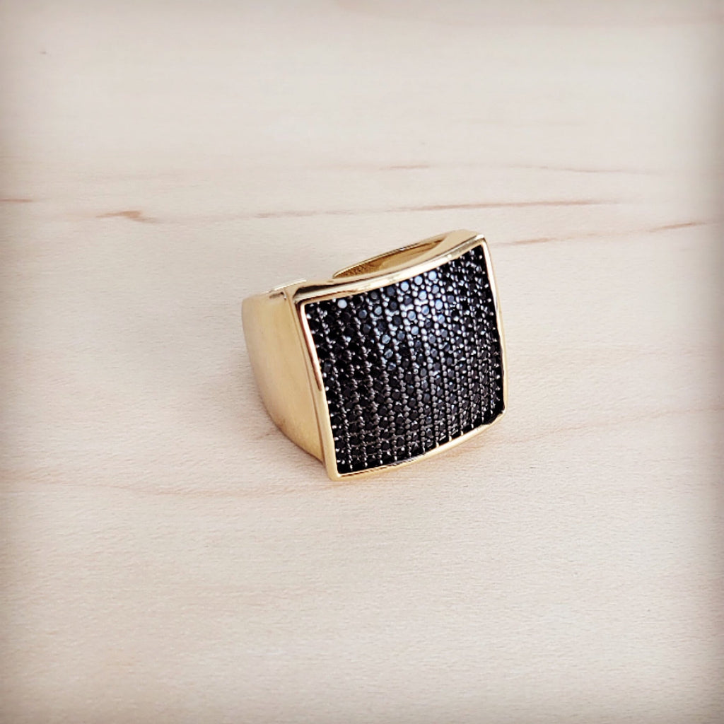 A gold-tone ring with a large square face completely set with black micro-pave crystals on a wooden background