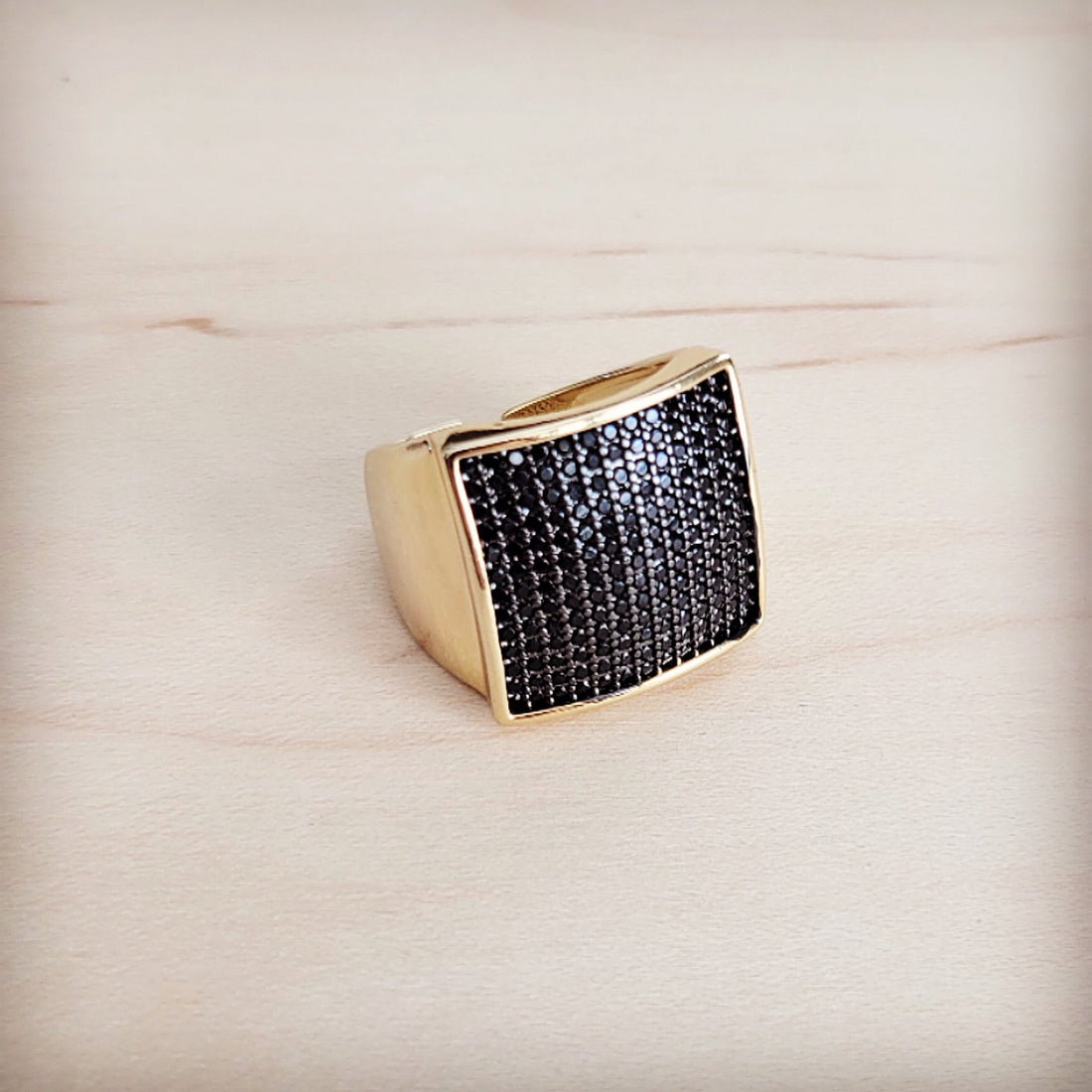 A gold-tone ring with a large square face completely set with black micro-pave crystals on a wooden background