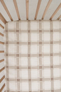 Top-down view of an Ellis Plaid muslin crib sheet in neutral cream and brown tones fitted on a wooden crib mattress.