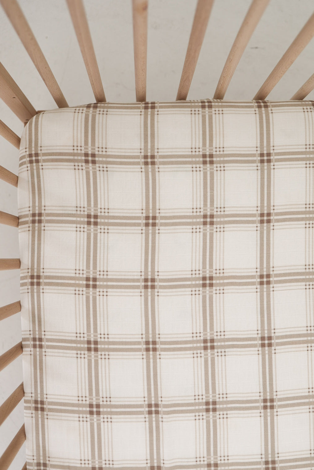 Top-down view of an Ellis Plaid muslin crib sheet in neutral cream and brown tones fitted on a wooden crib mattress.