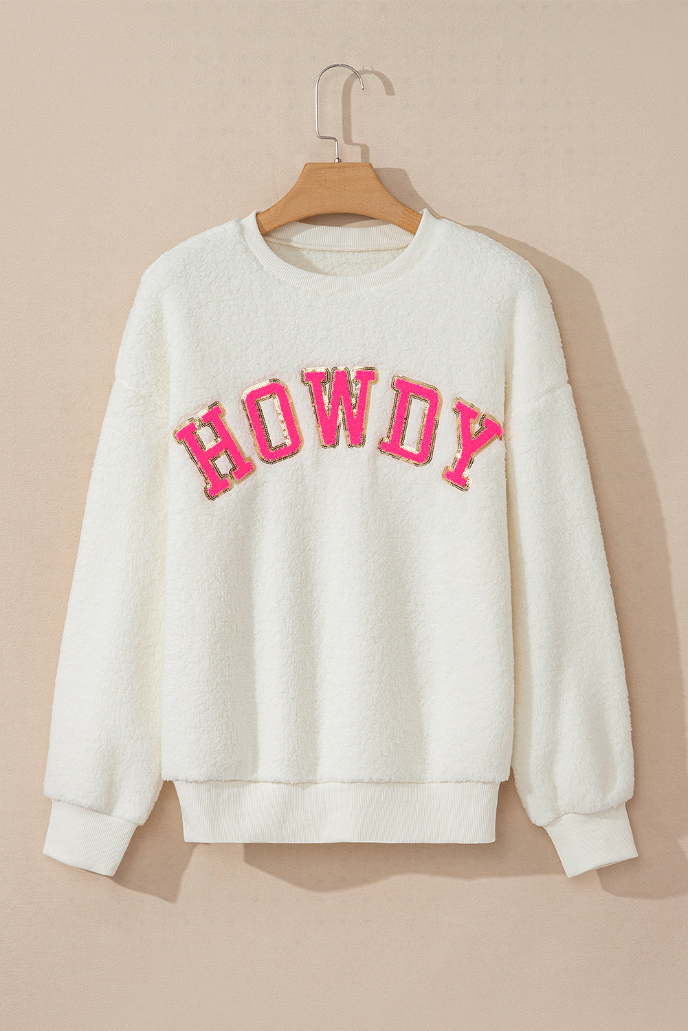 Front view of a white crew neck sherpa pullover featuring a pink and gold HOWDY letter patch on the chest, displayed on a wooden hanger against a neutral background.