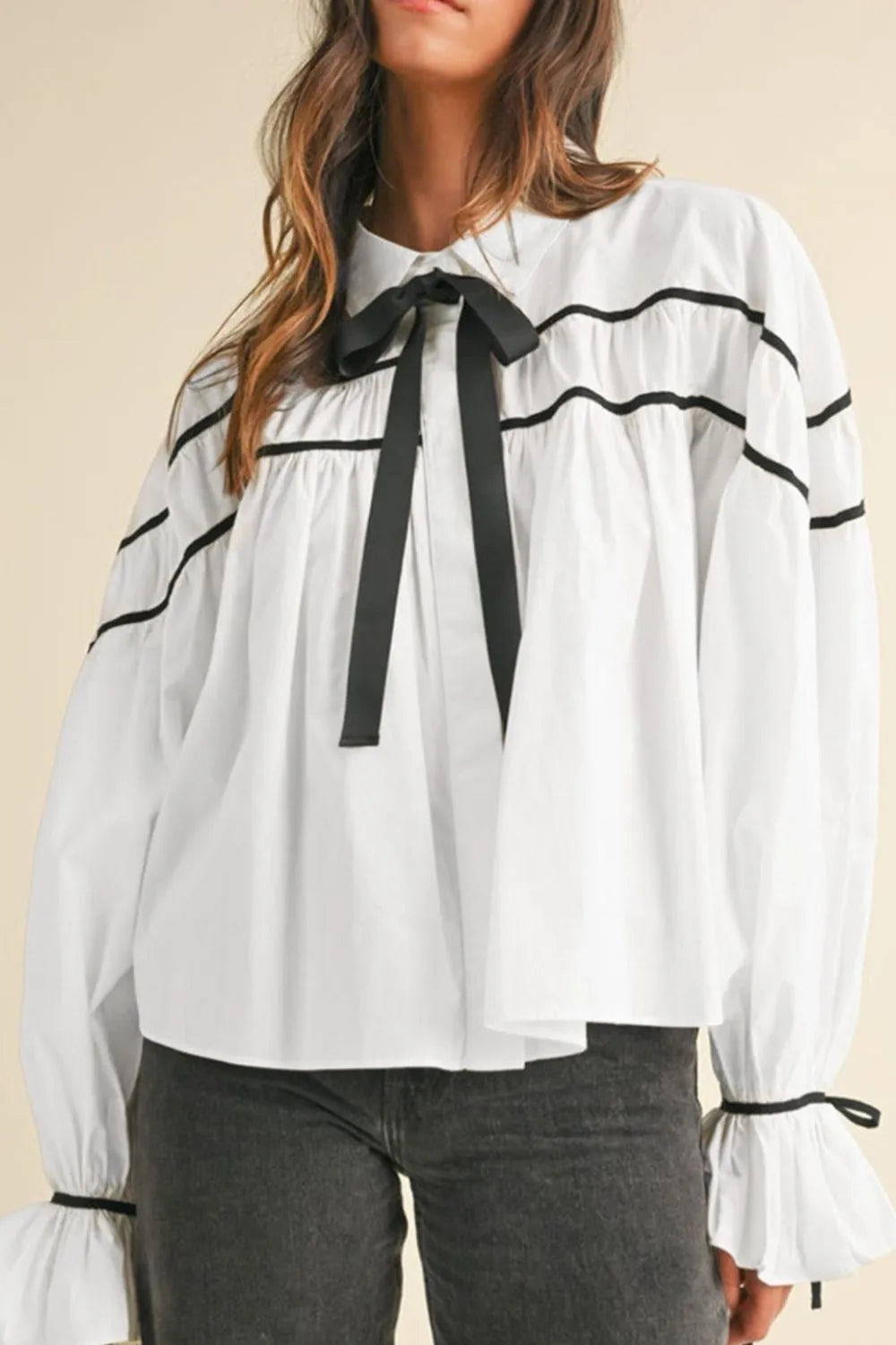 A woman wearing a white collared shirt with a black ribbon bowtie, featuring long flounce sleeves and black trim detail, styled with casual pants.