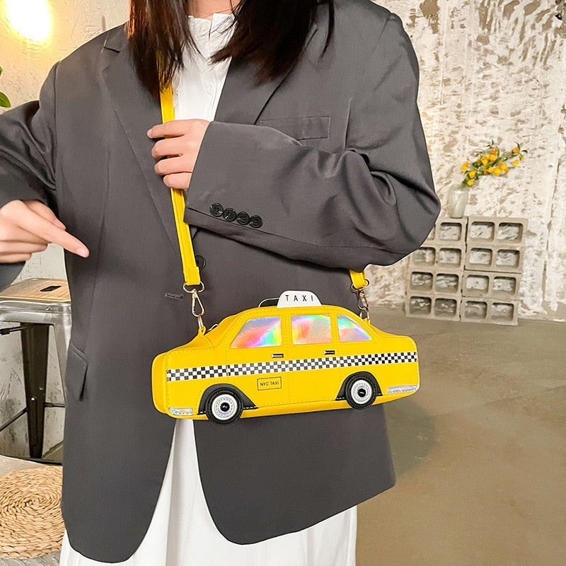 Vibrant yellow 3D taxi car shaped crossbody handbag featuring a black and white checkered stripe, iridescent windows, and 'NYC TAXI' detail, displayed on a person wearing a grey blazer.