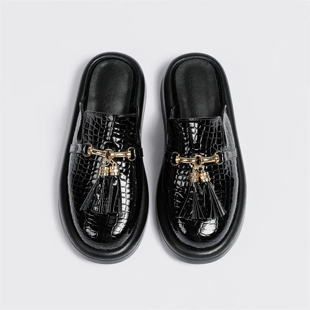 A pair of glossy black croc-embossed tassel mule flats with gold-tone hardware, shown from top-down and side views on a white background.
