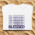 White crewneck t-shirt with a blue repeating BLESSED graphic print laid flat on a sandy surface.