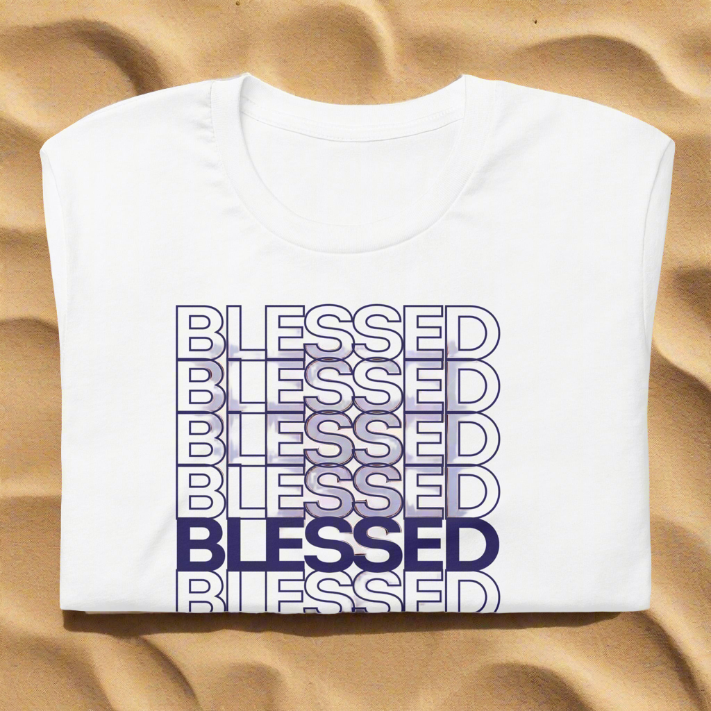 White crewneck t-shirt with a blue repeating BLESSED graphic print laid flat on a sandy surface.