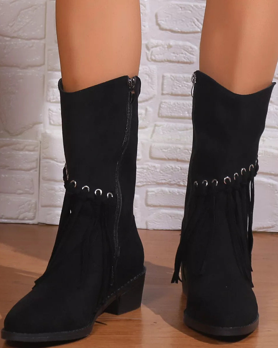 Black suede boots with fringes and decorative studs worn by a person against a white brick wall.