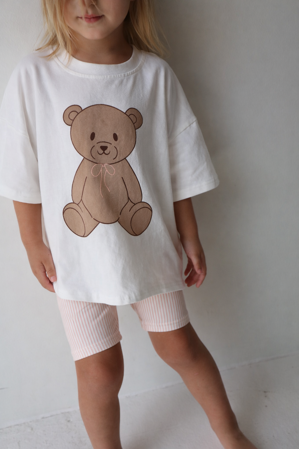A young girl wearing a white t-shirt with a brown teddy bear graphic and pink striped bike shorts.