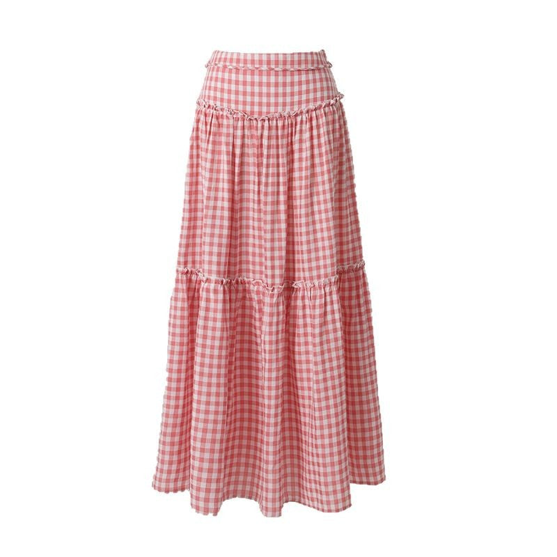 A woman models a pink and white plaid tiered maxi skirt with an A-line silhouette, paired with a white crop top and platform sandals. The skirt features delicate ruffle details on each tier. A flat lay image also shows the skirt clearly.