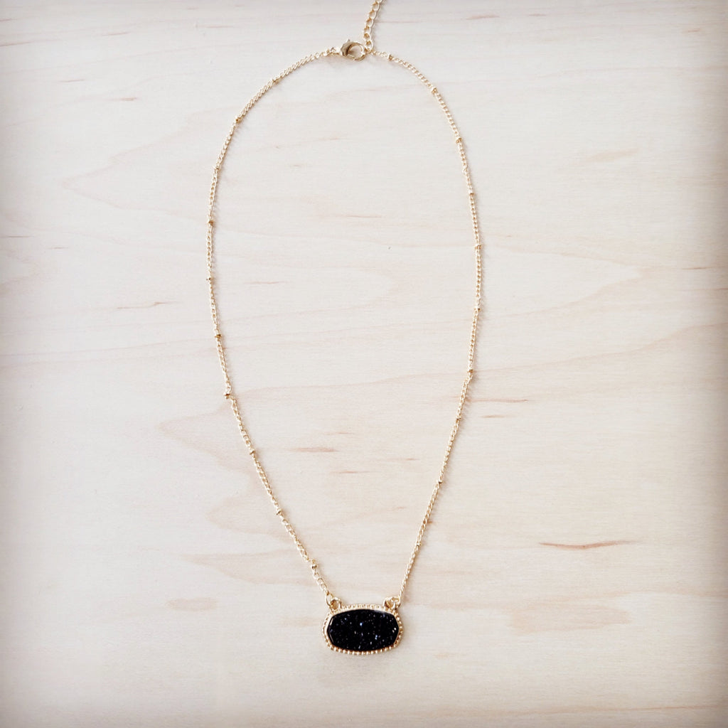 A gold-tone chain necklace with small metal knots and an oval black druzy pendant on a light wood background.