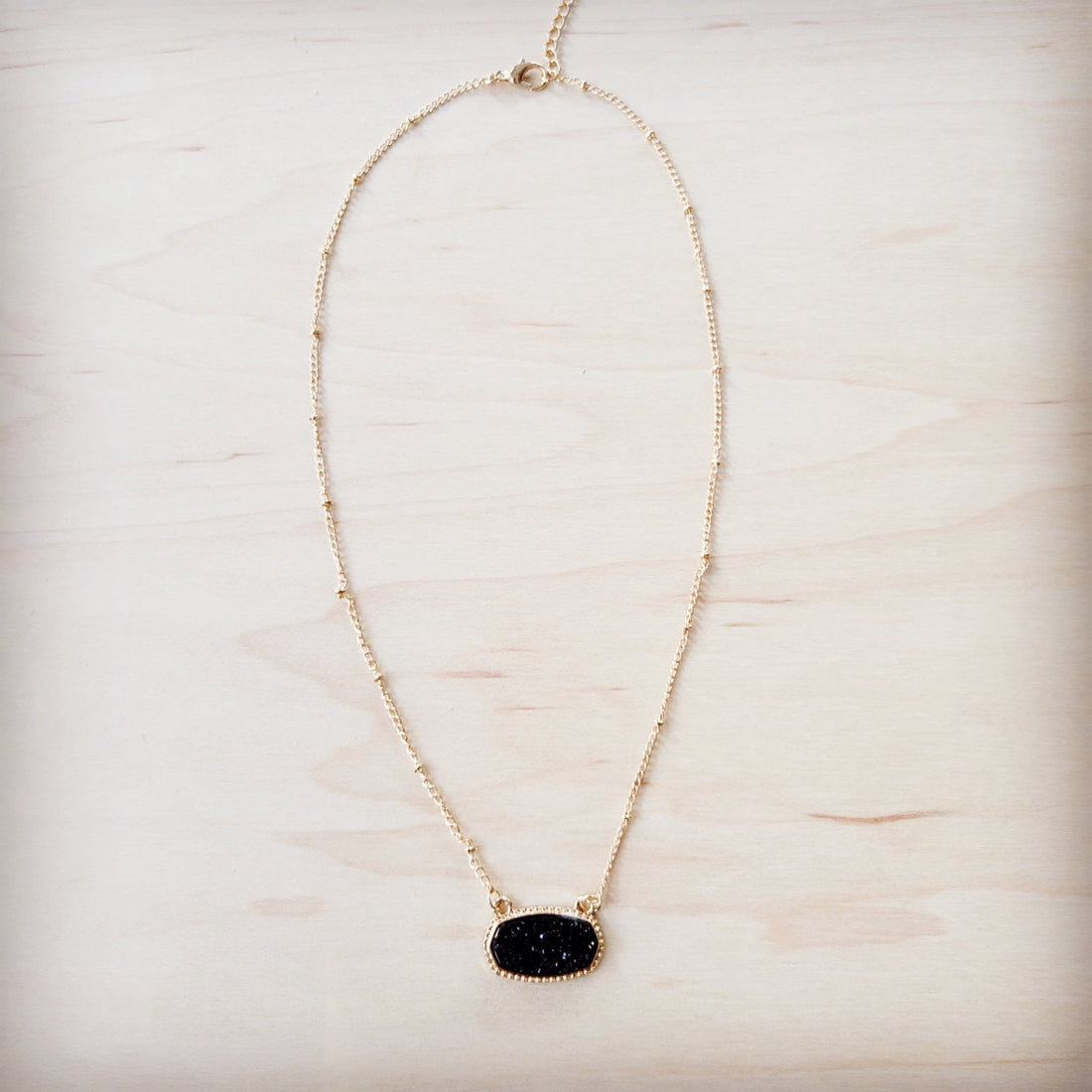 A gold-tone chain necklace with small metal knots and an oval black druzy pendant on a light wood background.